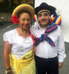Maria Mayda wears a red flower in her hair, standing next to a man with a multi-colored woven stash around this shoulder.