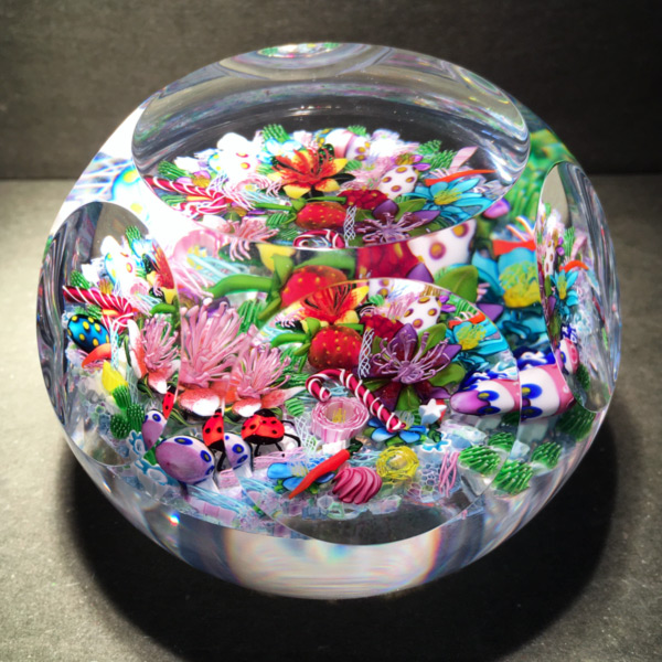 A Ken Rosenfeld Scramble Glass Paperweight with six cut windows. The view shows a medley of flamework objects, including a candy cane, lady bug, strawberry, and purple, pink, and blue flowers.