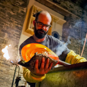 Headshot of Glass Artist David King, rolling a blowpipe with glowing hot glass. Smoke rises up from either side of the sheets of newspaper that he uses to shape the molten glass. Photo credit: Angus Mackay