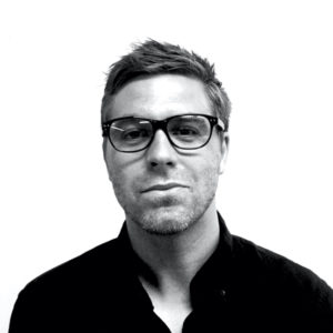 Black and white headshot of artist and educator David Schnuckel. He sports a short haircut and wears a dark shirt and glasses.