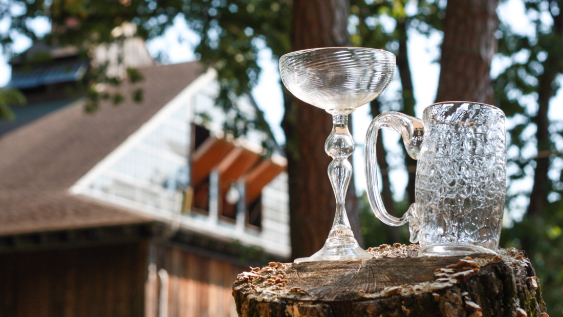 A glass wine and beer mug resting on a tree stump with a blurred view of the Glass Studio behind them.