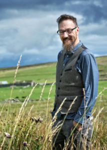 Artist Jeff Zimmer smiles in a field of wheat, a cloudy sky behind him. Photo Credit: Angus Mackay