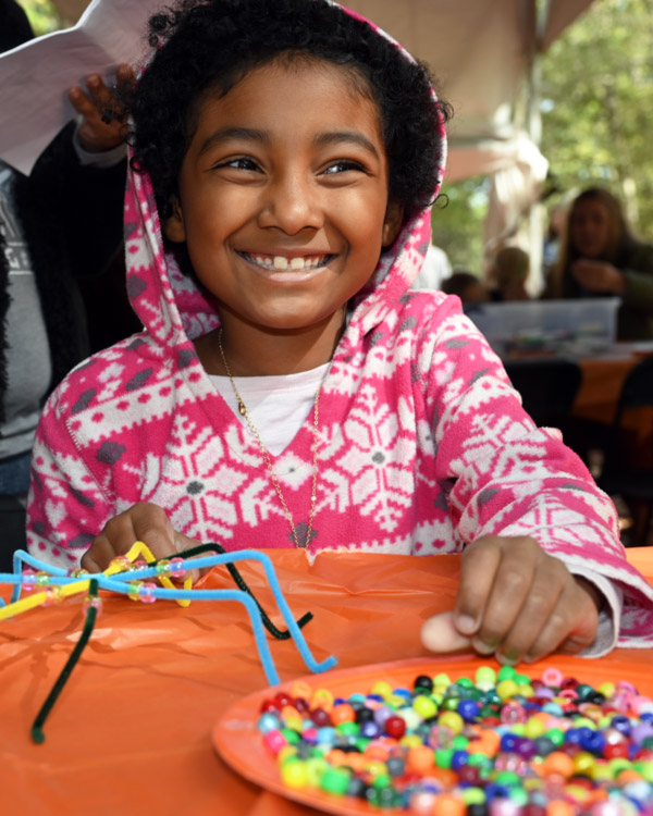 A child smiles wide as they work on a craft with pipe cleaners and beads.