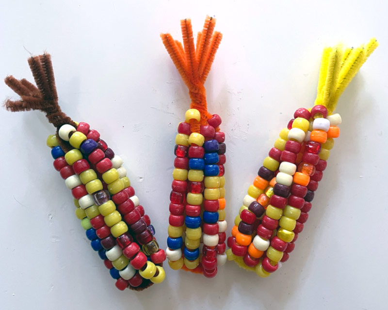 Three examples of the Beaded Fall Corn craft. The beads in red, yellow, white, and brown, wrap around brown, orange, and yellow pipe cleaners.