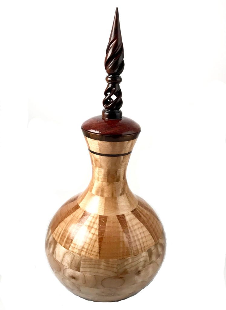 A tan wooden vessel with a lid by Scott Foster. The lid is dark brown with a carved topper coming up to a point.