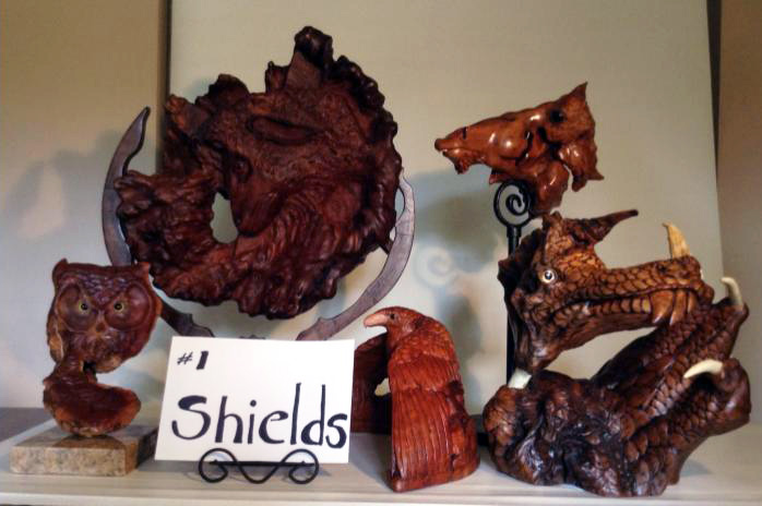 Several wood carvings by David & Barbara Shields. From left to right: a carved owl on a platform, a carved sheep in an organic circle shape, a small reddish brown hawk, and some carved heads of scaled animals.