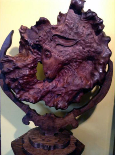 A wood carving in a deep magenta color of a sheep's face in an organic circle shape.