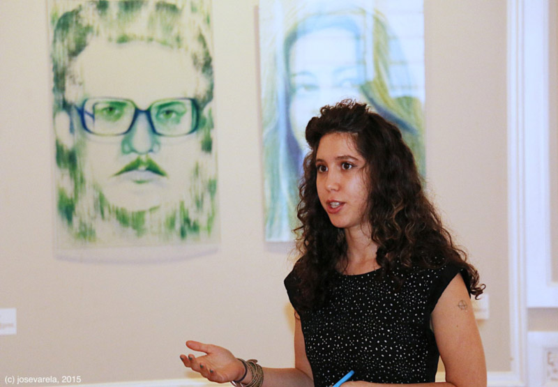 Paula Meninato speaks in front of glass panels with painted portraits. Photo by Jose Varela, 2015.