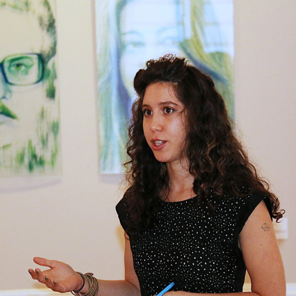 Paula Meninato speaks in front of glass panels with painted portraits. Photo by Jose Varela, 2015.