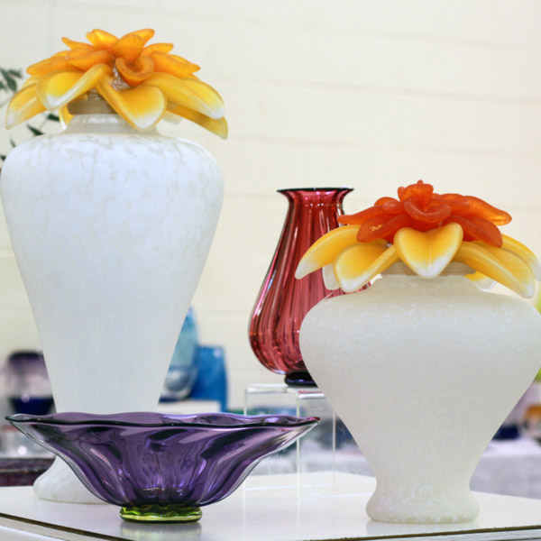 Two urns from Melanie and David Leppla. One is tall and frosted white with a vibrant yellow glass blown flower on top. Next to it is a stout, similar frosted white urn with a yellow and red glass flower on top.