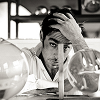 Marc Barreda has his head down with his hand on his forehead. He looks up at the camera. In the foreground are large glass spheres. Photo by Myscha Oreo.