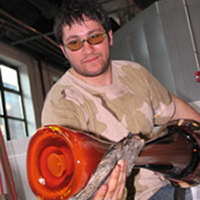 A photo of Moshe Bursuker from www.moshebursuker.com. Moshe shapes a piece of red hot glass in his studio.
