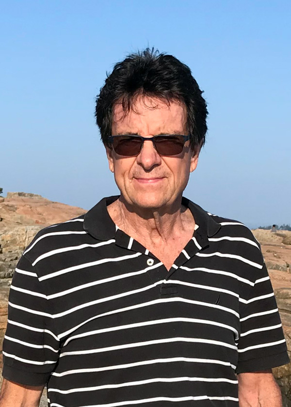 Dan Dailey stands outdoors. He has dark hair and wears brown sunglasses and a black and white striped button-up.