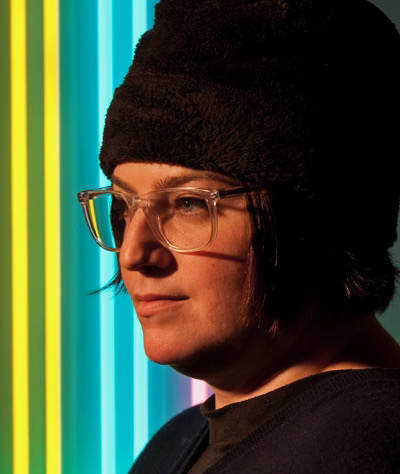 A headshot of artist Jen Elek. Jen is looking to the side with neon lights in the background. She wears clear glasses, a black top, and a black beanie.