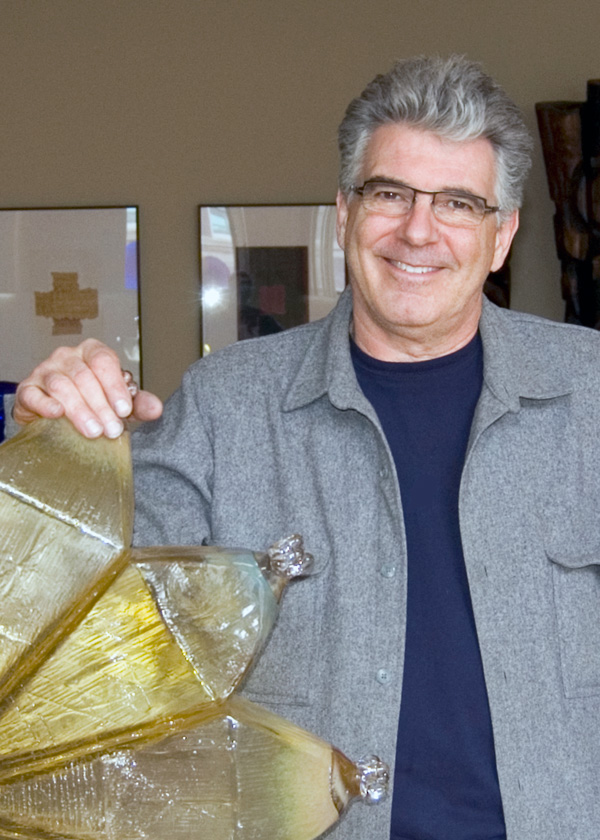 Glass Artist Richard Royal stands smiling, setting a hand on a piece of his work. He has grey hair and dark eyes and wears a gray blazer and a deep blue shirt.