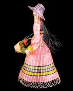 A horse hair miniature of a female figure in a purple hat and pink dress, carrying a basket of flowers.