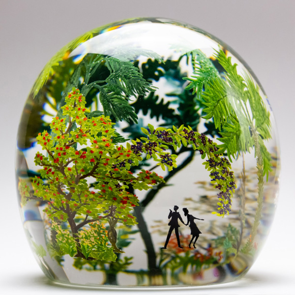 A paperweight shaped like a sphere with a flat bottom. On the outside, there are large painted leafy plants and trees. Inside there are the silhouettes of two people looking around into the jungle of plants.