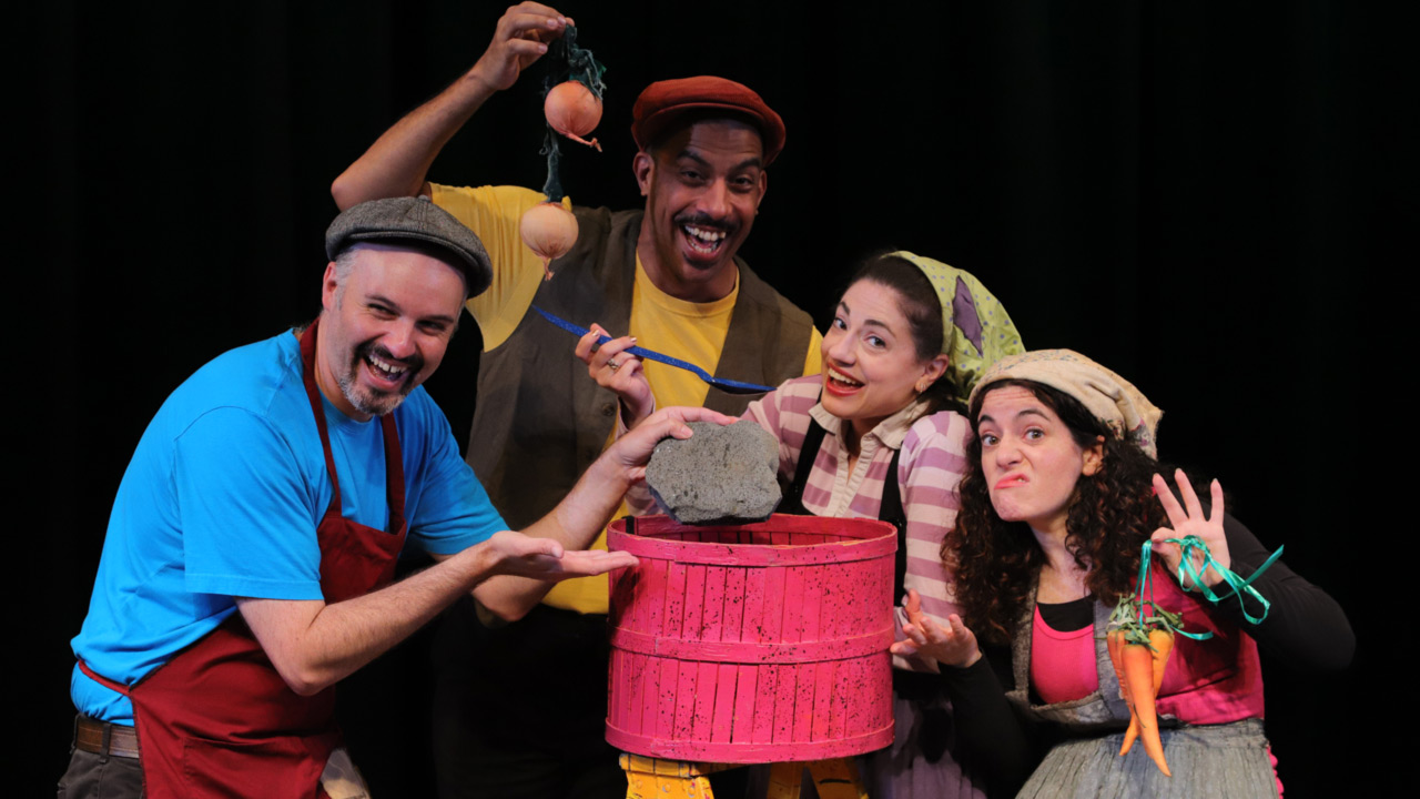 Four actors dressed in shabby clothes and aprons pose for a photo huddled over a circular make believe pot. There are two men who each wear mustaches and flat cap hats and hold items overtop the imaginary pot. Two women dressed in aprons and head scarves also hold veggies and utensils over pot.