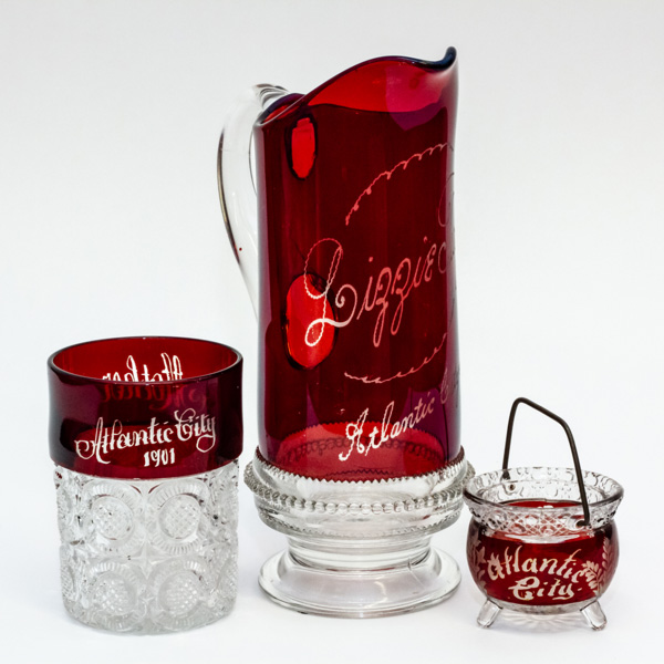 A small grouping of red glass with "Atlantic City" written in white on each. A small clear cut glass cup with a thick translucent red rim. In the middle, is a tall red pitcher with 'Lizzie' written on it and a cut glass base, and lastly a small cut glass vessel with a red body.