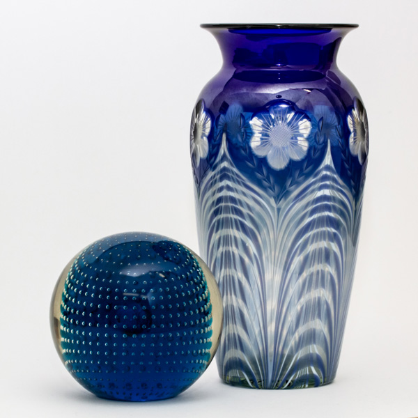 A deep blue sphere paperweight with small clear dots in rings around the sphere from top to bottom. Next to the paperweight is a tall vase in a similar blue with tall white leaves and flowers in between near the wide lip of the vase.