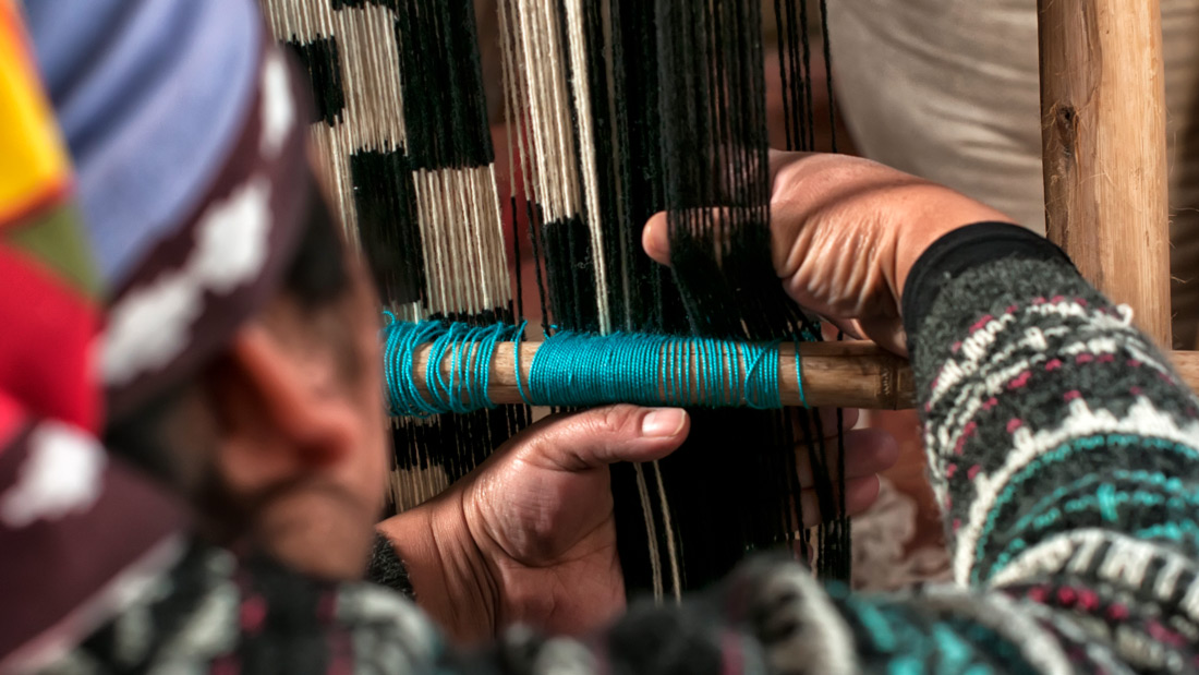 A textile designer gathers colorful yarn in their hands and weaves them into a pattern. The yarn hangs over a wooden loom. The designer stands in front of the loom while they work.