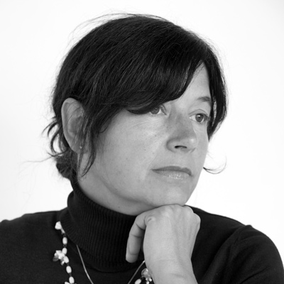 In black and white, artist Syblle Peretti poses for a headshot in front of a white background. Syblle wears a black turtleneck with two necklaces: one a single silver chain and the second a loose string of pearls. Syblle looks off to the right of the frame and rests her chin on her knuckles. Syblle wears a pixie haircut and small pearl earrings.