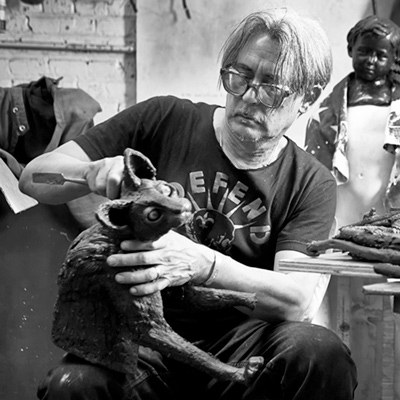 A black and white photo of artist Stephen Paul Day who carves a wooden figure of a “fox”. Stephen sits in the middle of a white bricked room with a dark childlike figure sitting behind him on his left. There is a work table in front of Stephen to his left with various carving tools piled up. A dark windbreaker jacket is tossed over the back of a covered chair behind Stephen and on his right. Stephen wears a dark t-shirt with the words “DEFEND” written in white text arched over a circular graphic on the front and black jeans.