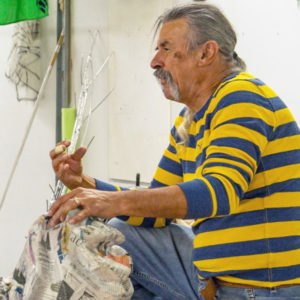 Peruvian artist Gustavo Boada stands in front of a white wall in a raised-leg pose as he speaks to an unknown audience off frame.