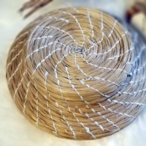 Pine Needle basket by artist Tyrese Gould Jacinto with light brown pine needles and white string arranged in a swirl pattern of the basket. The basket sits on a beige tablecloth with dark objects mostly out of frame and out of focus.