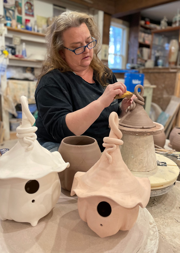 Pottery artist Phyllis Seidner sponges water onto a damp sculpture. Several pots at different stages of drying sit on a work table in front of Phyllis. A blurred out pottery workshop and full shelves in the background.
