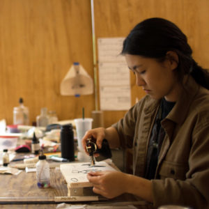 Artist Suyeon Kim sits at a work table and uses a small flame torch against a piece of wood. Multiple tools and items on a wooden table blurred in the background.