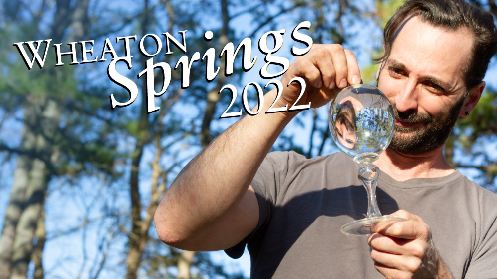 Glass artist Alex Rosenburg looks through an optical glass instrument with his reflection showing through to the other side of the glass. The trees and sky are blurred in the background. White text overlays that reads: “Wheaton Springs 2022”.