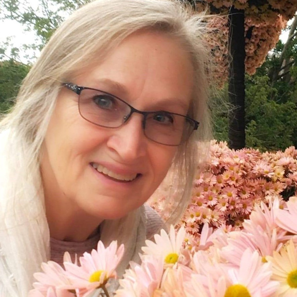 Veronica Kairos poses for a headshot outdoors. Veronica poses next to pink flowers with a tree and several bushes of pink flowers in the background. Veronica wheres black glasses, a light-colored shirt, and a light-colored scarf.