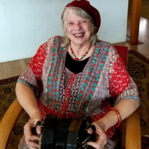 American country music artist Lisa Stewart Garrison poses for a headshot as she sits in a chair in the middle of a small room. Lisa holds a musical instrument in her lap.