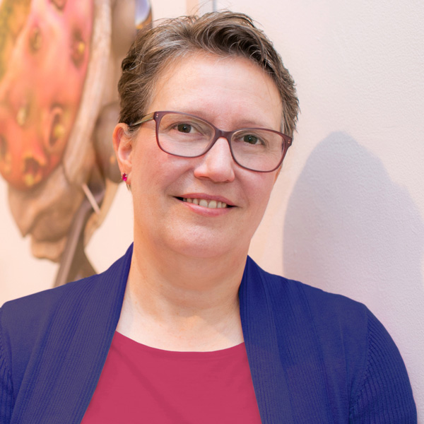 M. Rothwell poses for a closeup in front of a white wall and wears a navy blue cardigan, dark pink shirt, brown glasses, and a close-cut short haircut.