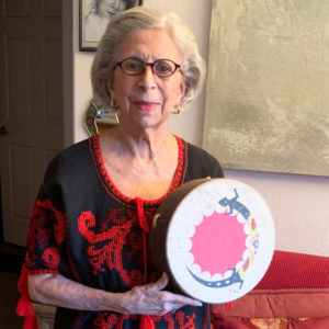 Argentinian folk musician Suni Paz poses for a headshot in a small room in front of a red couch. She holds a decorative musical instrument and a red and black dress.