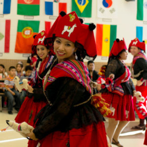 Boliviana Diablada Llamerada Dance dancers perform for a room of school-aged children. There are flags of multiple countries displayed on a wall in the background.