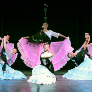 Elenco Paraguay Danza performers pose on a stage in pink and blue festive skirts, white lace tops, and black cultural shawl wraps.
