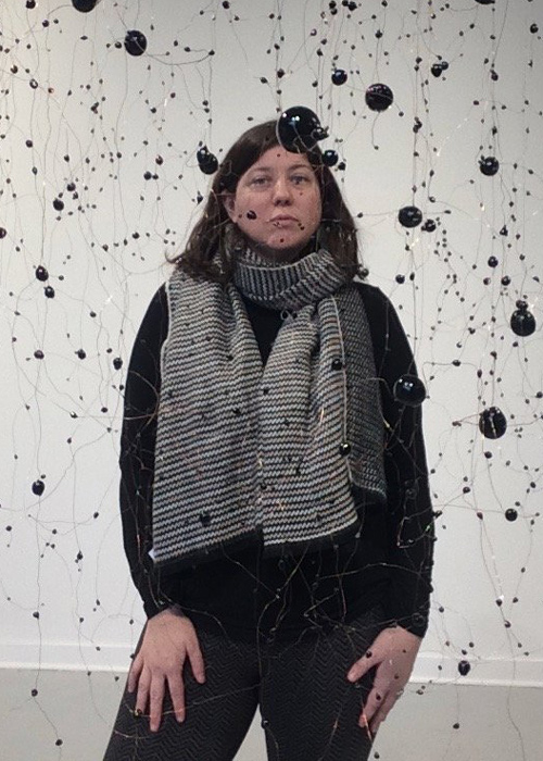 Jessica Julius poses for a headshot in the middle of a hanging display of black orbs. Jessica wears a black sweater, darks pants, and a black and white stripe scarf.
