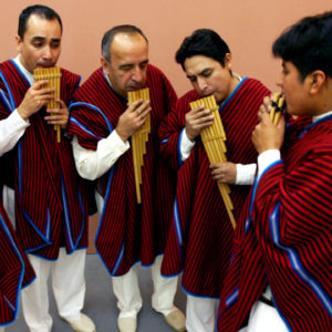 Pepe Santana Inkhay Anean Music performers play wood instruments as they wear cultural red and white clothing. They stand in front of a peach-colored wall.