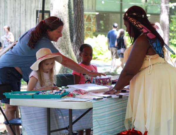 Fantasy Faire 2019 attendees participate in kids' craft activities with a Fantasy Faire actor dressed in a whimsical yellow dress and colorful butterfly wings at a tented booth outdoors. Other attendees walk passed in the background.