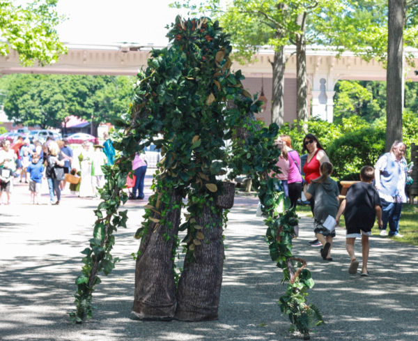 Cropped image of Fantasy Faire 2019 attendees and actors walk past and admire a life-like tree creature along the WheatonArts campus outdoors.