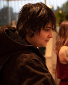 Headshot of visual artist Kayla Cantu wearing a short haircut, lower lip piercing, and black hooded coat. There are trees and another person wearing long brown hair and a red dress out of focus in the background.