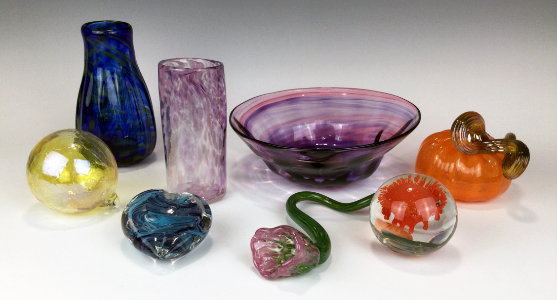Several Make-Your-Own groupings of colorful hot glass bowls, glasses, vases, and paperweights sit arranged on a white display space.