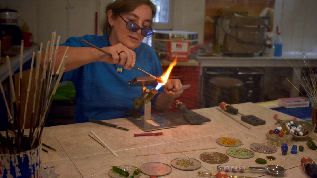 Flamework artist is creating glass beads with a torch tool in the center of the image. In front of her are: a cup of paintbrushes, an assortment of tools, and small trays containing different colored beads. There are orange, green, brown, and pink. The artist is wearing a blue shirt and protective goggles. She is working the ceramics studio.