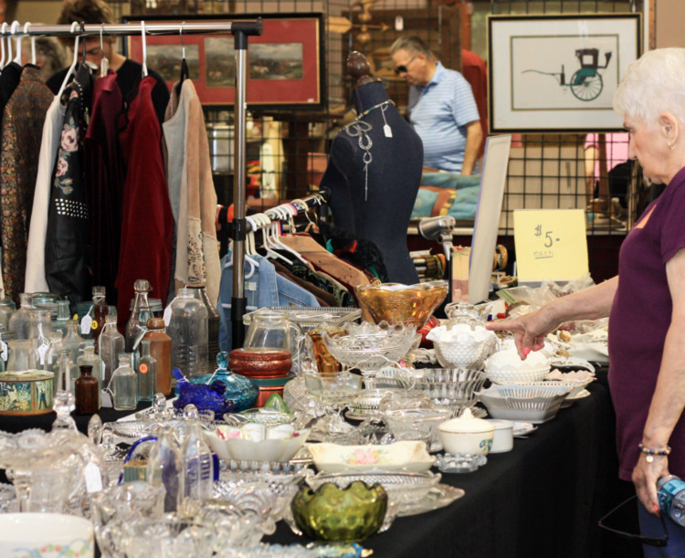 A woman in a purple shirt on the right side of the image is pointing at a glass dish at an antique vendor's table in the vent hall. The subject is the table with an array of antique dishes labeled $5 each. In the background is an antique clothing vendor and a man in a blue polo browsing an unseen vendor. the man is standing between two framed pictures. The left picture has a red bored and the subject is a farm landscape. The right picture is of a teal and black carriage attachment.