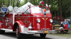 Red fire truck on the WheatonArts grounds for the annual fire muster event. A man in a blue shirt is driving the vehicle. Another man in a blue shirt is observing the festivities from the back of the truck. To the right of the truck is a family with kids sitting at a picnic table, watching the truck go by.