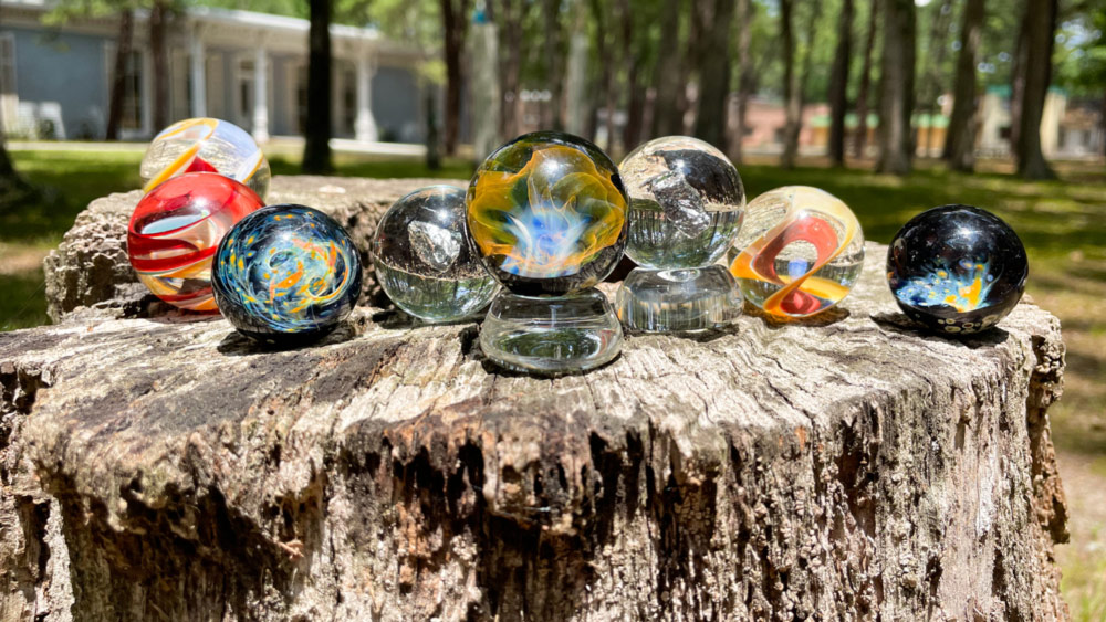 The subject of the image is an array of multicolored glass paperweights centered in the image on a stump with the Museum of American Glass in the background. There are eight paperweights each with their own layered designs. They are all space themed and look as if there are tiny galaxies in them.