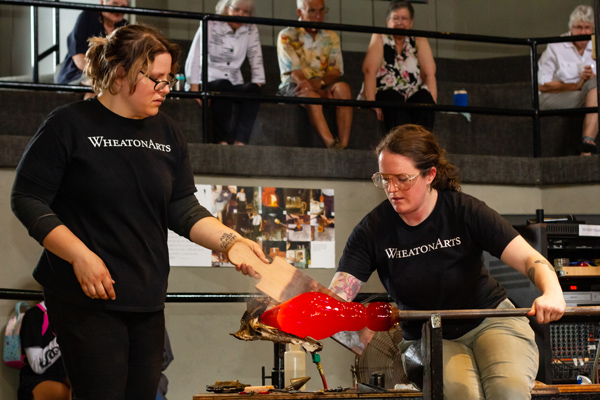 Tiffany and Katie, two glass artists, are shaping a glass piece on the studio floor in the center of the image. Tiffany, the artist on the left, is holding a wooden paddle to control the edges of the glass while Katie on the right is turning the piece on a long metal tool. At the top of the image is five visitors watching the process from above.