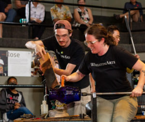 Two glass artists are in the process of creating a glass piece in the studio. On the left, one artist is holding what looks like to be a lank of wood up to the piece while the other artist on the right is holding the rod the piece is attatched to and a glass handle. The piece is a deep blue, almost purple, vase with gold handle. In the background are several people observing the process.